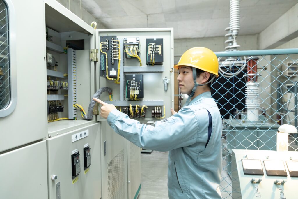 九州電気専門学校の電気設備実習風景。ヘルメットと作業着を着用して受電盤を確認する様子